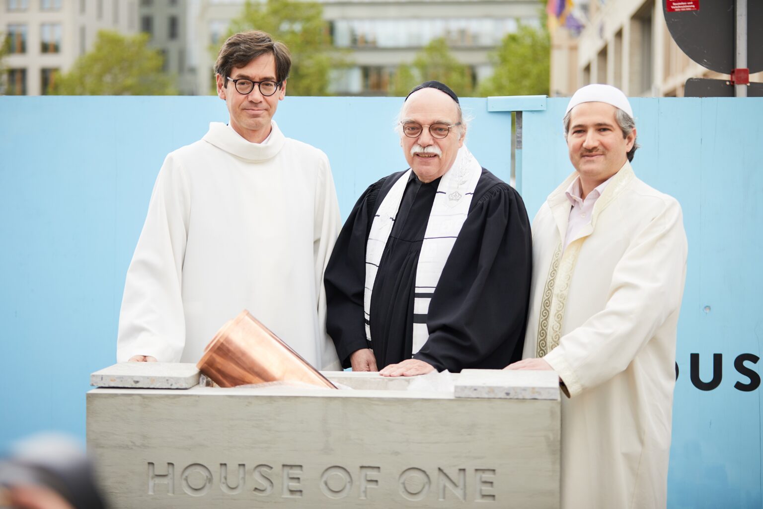 Ein christlicher Priester, ein jüdischer Rabbiner und ein muslimischer Imam stehen gemeinsam vor dem Grundstein des House of One in Berlin. Sie symbolisieren den interreligiösen Dialog und das gemeinsame Ziel, einen Raum für Frieden und Verständigung zu schaffen. Im Hintergrund ist eine blaue Bauwand sowie ein urbaner Gebäudekomplex zu sehen.